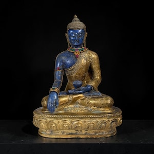 Peut inclure: Une statue de Bouddha assise avec une finition bleu profond sur la tête, le torse et les bras, contrastant avec une robe dorée. La figure porte un collier avec des pierres vertes et rouges, tient un petit bol sur les genoux et repose sur une base lotus.