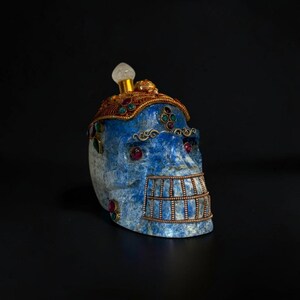 May include: A blue lapis lazuli skull sculpture adorned with gold-toned metal filigree, red and green cabochon gemstones, and a clear crystal topper. The jaw area features a gold-toned metal cage overlay with a grid pattern across the teeth.