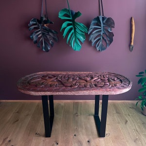 Könnte beinhalten: Ein ovaler Tisch aus geschnitztem Holz mit aufwendigen Designs. Der Tisch hat schwarze Metallbeine und steht auf einem Holzboden. Der Hintergrund ist eine dunkelviolette Wand mit drei grünen und schwarzen Blattdekorationen, die von der Decke hängen.
