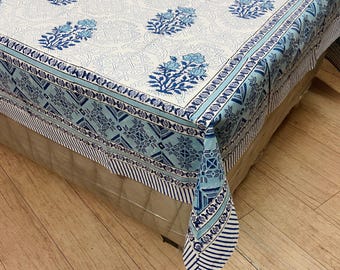 May include: A white and blue floral patterned tablecloth. The tablecloth features a repeating pattern of blue floral bouquets and geometric designs. The border has a contrasting design of stripes and geometric shapes. The tablecloth is draped over a surface.