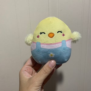 Puede incluir: Un peque&ntilde;o juguete de peluche amarillo en forma de pollito con un mono azul y una flor blanca. El pollito tiene ojos bordados y un pico rosa.
