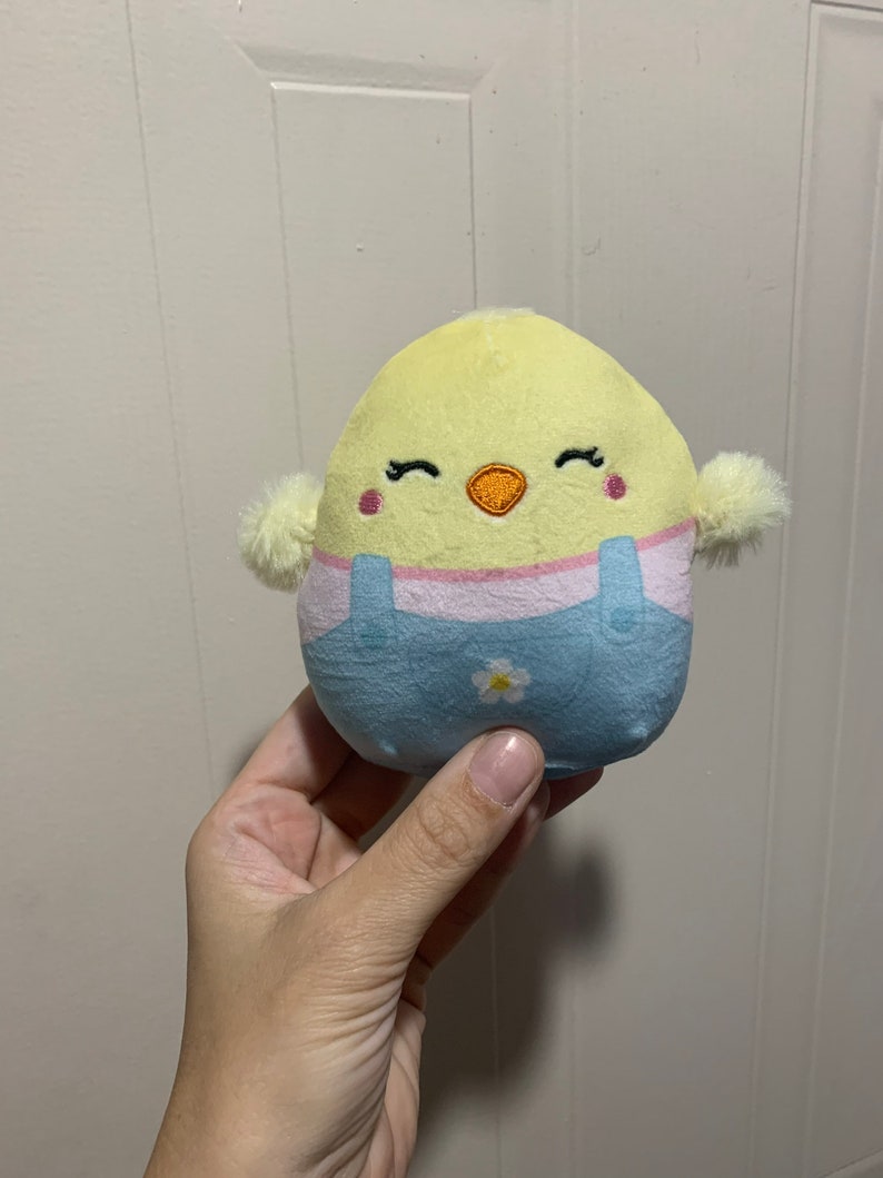 Puede incluir: Un peque&ntilde;o juguete de peluche amarillo en forma de pollito con un mono azul y una flor blanca. El pollito tiene ojos bordados y un pico rosa.