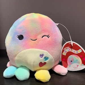 Può includere: Un giocattolo di peluche a forma di polpo, con un motivo tie-dye arcobaleno, un viso che fa l'occhiolino e cuori sul ventre. Il giocattolo è etichettato "Original Squishmallows" e "Opal".