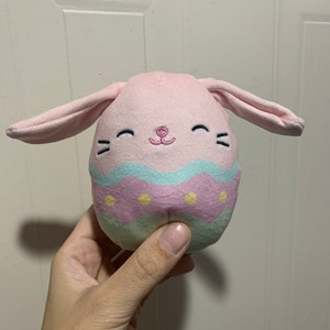 Puede incluir: Un juguete de peluche rosa en forma de huevo de Pascua con orejas de conejo. El huevo est&aacute; decorado con rayas azul pastel, rosa y verde y puntos amarillos. El conejo tiene una nariz rosa y ojos bordados.