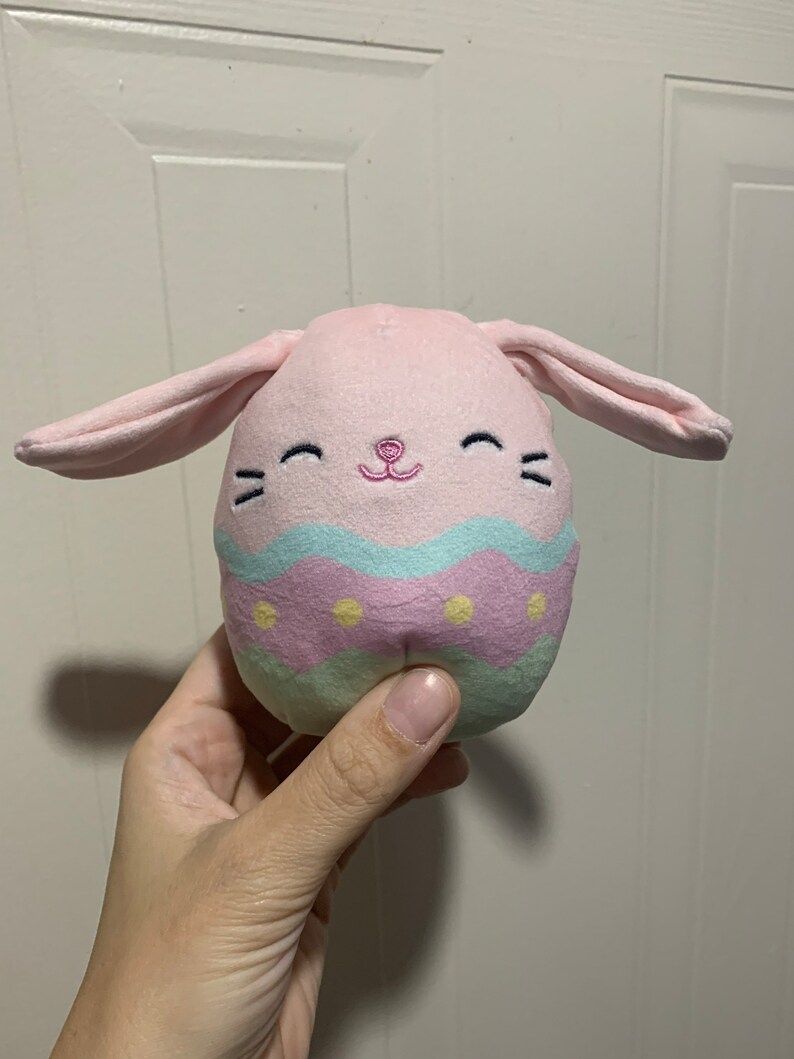 Puede incluir: Un juguete de peluche rosa en forma de huevo de Pascua con orejas de conejo. El huevo est&aacute; decorado con rayas azul pastel, rosa y verde y puntos amarillos. El conejo tiene una nariz rosa y ojos bordados.