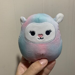Puede incluir: Un peque&ntilde;o juguete de peluche con forma de oveja con un patr&oacute;n tie-dye en tonos azules, rosas y blancos. La oveja tiene una cara blanca con ojos negros bordados, pesta&ntilde;as y una nariz rosa.