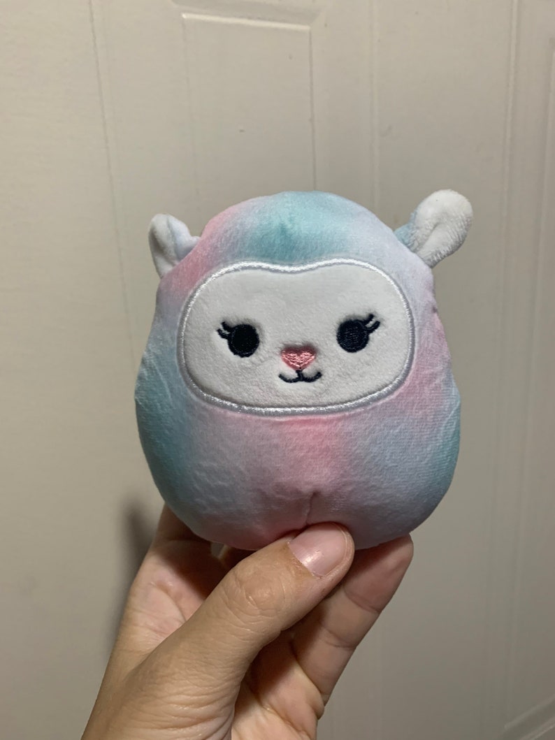 Puede incluir: Un peque&ntilde;o juguete de peluche con forma de oveja con un patr&oacute;n tie-dye en tonos azules, rosas y blancos. La oveja tiene una cara blanca con ojos negros bordados, pesta&ntilde;as y una nariz rosa.