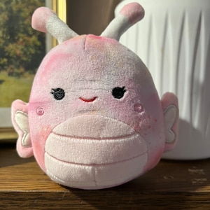 Puede incluir: Un juguete de peluche rosa y blanco con forma de mariposa con una cara sonriente. El juguete tiene una textura suave y peluda y est&aacute; hecho de un material de alta calidad.