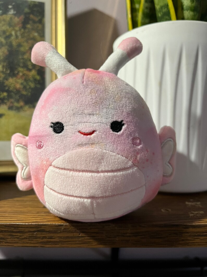 Puede incluir: Un juguete de peluche rosa y blanco con forma de mariposa con una cara sonriente. El juguete tiene una textura suave y peluda y est&aacute; hecho de un material de alta calidad.