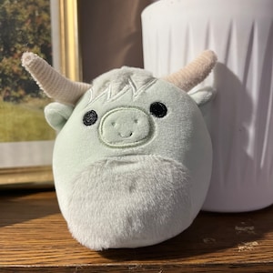 Puede incluir: Un juguete de peluche con forma de vaca con un cuerpo verde claro, cuernos blancos y ojos negros. El juguete tiene una sonrisa cosida y una textura peluda.
