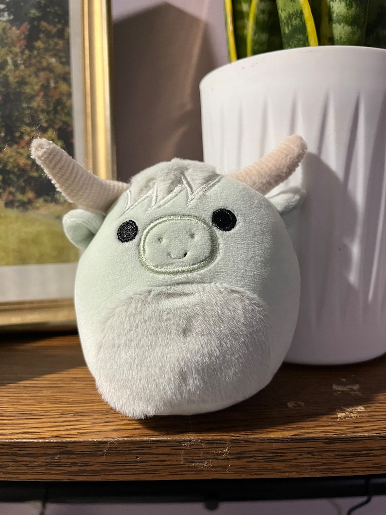 Puede incluir: Un juguete de peluche con forma de vaca con un cuerpo verde claro, cuernos blancos y ojos negros. El juguete tiene una sonrisa cosida y una textura peluda.
