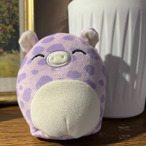 Puede incluir: Un cerdo de peluche morado con manchas blancas y una barriga blanca. El cerdo tiene una sonrisa cosida y un hocico blanco.