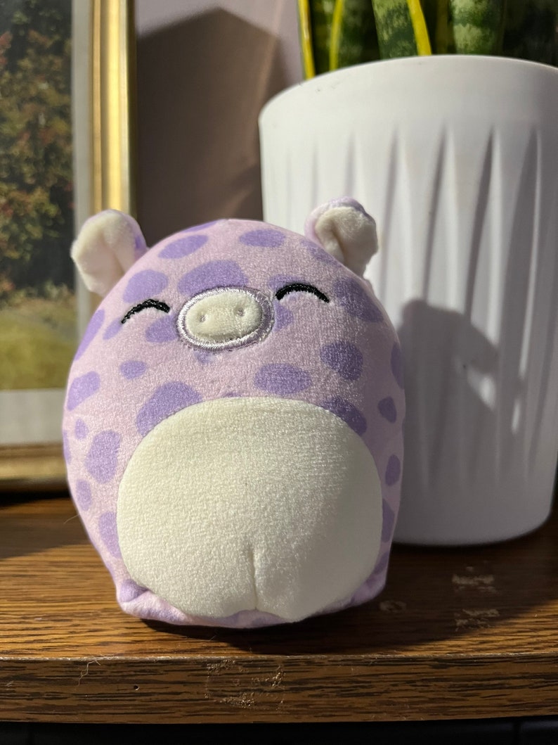 Puede incluir: Un cerdo de peluche morado con manchas blancas y una barriga blanca. El cerdo tiene una sonrisa cosida y un hocico blanco.