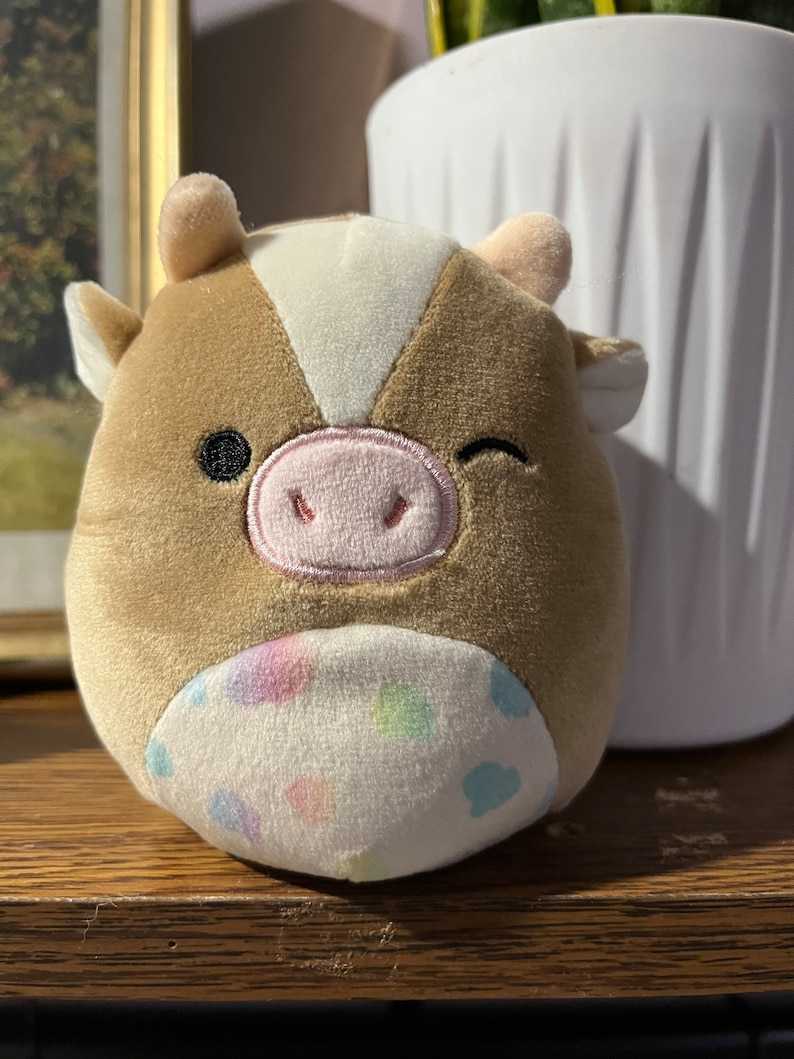 Puede incluir: Una vaca de peluche marr&oacute;n y blanca con la nariz rosa y un ojo gui&ntilde;ando. La vaca tiene un cuerpo blanco con lunares de colores.