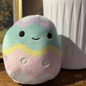 Puede incluir: Un juguete de peluche con forma de huevo de Pascua con un patr&oacute;n de rayas verde pastel, amarillo y rosa. El huevo tiene una cara sonriente con ojos negros y un contorno blanco. El huevo tiene lunares blancos en la secci&oacute;n rosa.