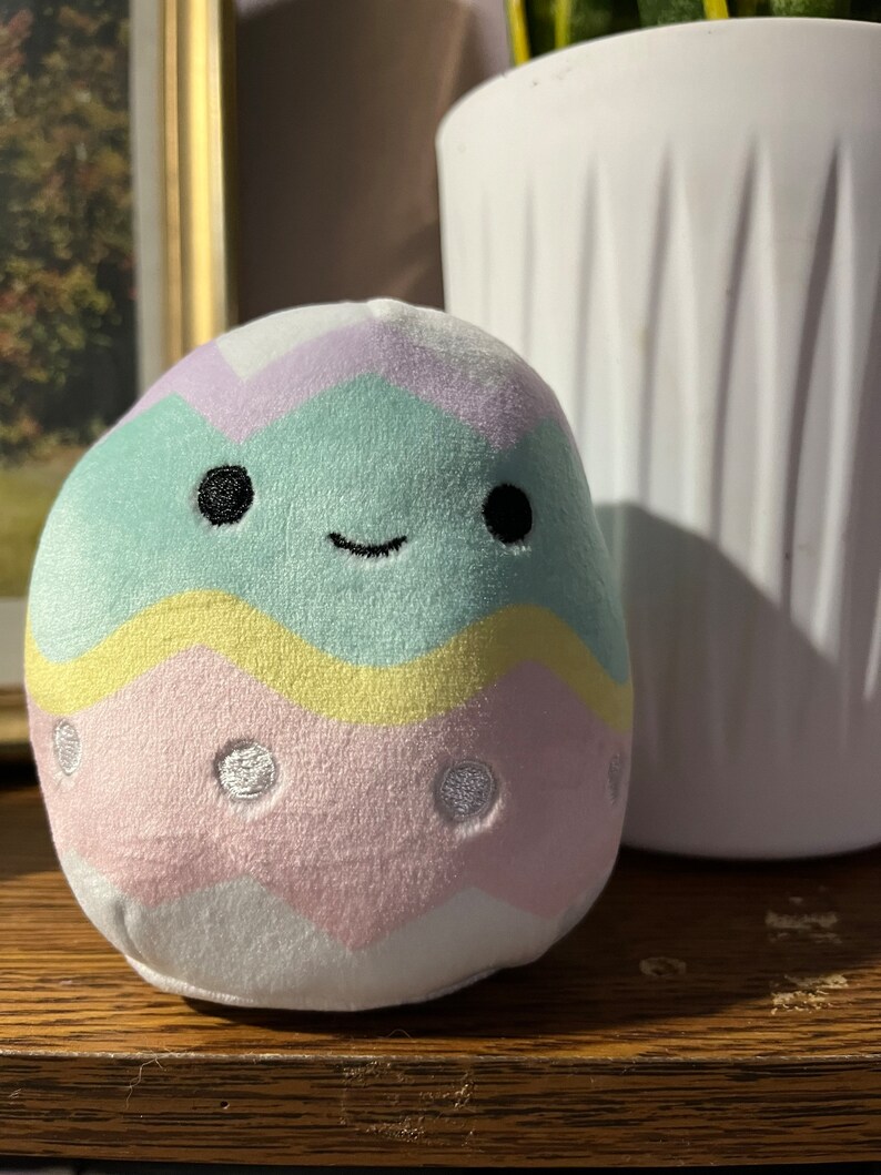 Puede incluir: Un juguete de peluche con forma de huevo de Pascua con un patr&oacute;n de rayas verde pastel, amarillo y rosa. El huevo tiene una cara sonriente con ojos negros y un contorno blanco. El huevo tiene lunares blancos en la secci&oacute;n rosa.