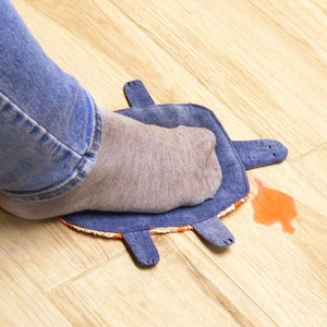May include: A blue denim turtle-shaped floor mat with four flipper-like appendages. A gray socked foot rests on the mat. An orange liquid stain is next to the mat on a light wood floor.