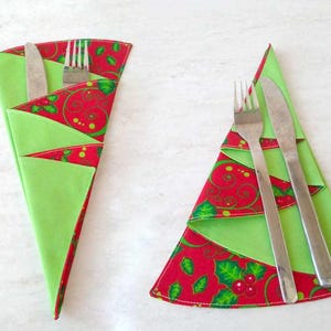 Puede incluir: Dos servilleteros con forma de árbol de Navidad con cubiertos. Los soportes son verdes con detalles estampados rojos con acebo y bayas. Un soporte contiene un tenedor y un cuchillo, el otro un tenedor. Decoración festiva navideña.