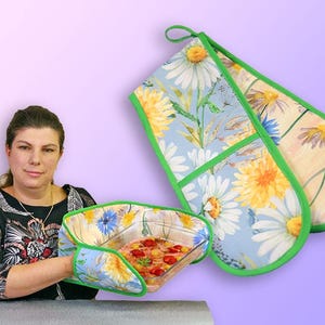 May include: Floral oven mitts and pot holder set. The mitts and pot holder feature a daisy pattern with yellow and white flowers on a light blue background, trimmed with green. The pot holder is holding a glass dish with food.