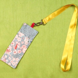 May include: A floral fabric lanyard with a yellow satin strap. The fabric pouch features a grey background with pink and red flowers. The lanyard is attached to a silver clasp and a red fabric piece.
