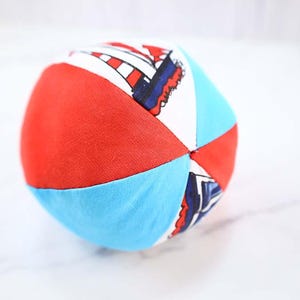 May include: A colorful, soft fabric ball with red, light blue, and white panels. Some panels feature a pattern with red and blue elements. The ball is designed for play and has a rounded shape.