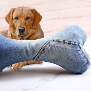 May include: A golden Labrador Retriever gazes at the camera, with a denim bone-shaped dog toy in the foreground. The toy is made of blue denim fabric with visible stitching. The dog is wearing a collar.