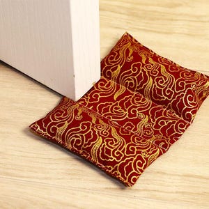 May include: Red and gold decorative door stop with a cloud pattern. The door stop is made of fabric and is rectangular in shape. It is placed on a light wood floor next to a white door.