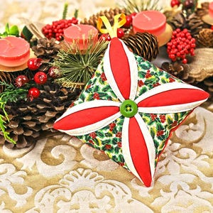 Pode incluir: Enfeite de Natal feito à mão com um design de estrela vermelho e branco. O enfeite apresenta um botão verde no centro e é decorado com um padrão de bagas de azevinho. O fundo inclui pinhas, velas e vegetação.