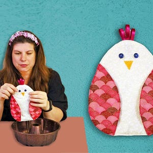 Puede incluir: Un guante de cocina acolchado blanco y rojo con forma de pollo, con ojos de botón azules y un pico amarillo. El guante tiene un detalle de cinta rosa en la parte superior. El fondo es de color turquesa.