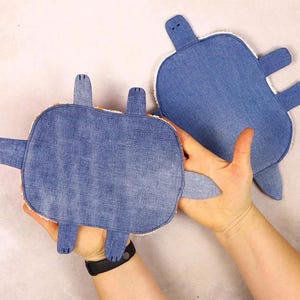 May include: Two denim turtle-shaped plush toys. The turtles have blue denim tops, white trim, and protruding flipper and head shapes. One turtle is held in a person's hand, while the other is on a light-colored surface.