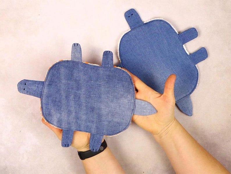 May include: Two denim turtle-shaped plush toys. The turtles have blue denim tops, white trim, and protruding flipper and head shapes. One turtle is held in a person's hand, while the other is on a light-colored surface.