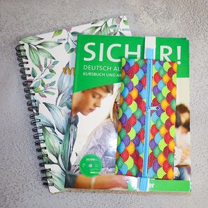 以下が含まれることがあります： 植物柄のスパイラルノートと、白で「SICHER！」の文字が入った緑色の本。鱗模様のカラフルなジッパー付きポーチが本の上にあります。