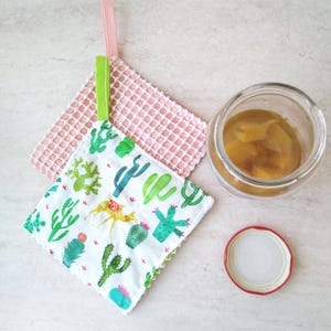 May include: Two square pot holders with hanging loops and a jar of preserves. One pot holder is white with a cactus print, the other is pink with a waffle pattern. The jar has a red-rimmed lid.