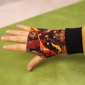 Może przedstawiać: Bezpalcowy mankiet na nadgarstek z czarnym mankietem i kolorowym wzorem pomarańczowych, żółtych i brązowych liści oraz kształtów przypominających żyrafy. Mankiety są noszone na dłoni, z widocznym żółtym rękawem.