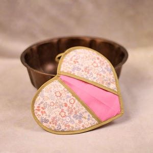 Könnte beinhalten: Ein herzförmiger Topflappen mit floralem Muster, rosa Akzent und goldfarbener Umrandung. Der Topflappen ist aus Stoff und hat eine Aufhängeschlaufe. Im Hintergrund befindet sich eine dunkelbraune Gugelhupfform.