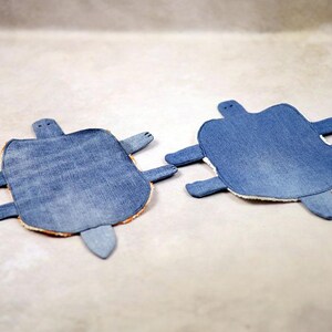 May include: Two denim turtle-shaped coasters. Each coaster features a blue denim top, a tan border, and denim appendages. The coasters are designed to resemble turtles, with a head, four flipper-like legs, and a tail.