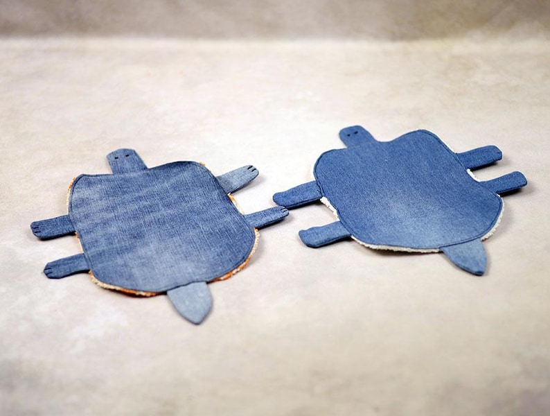 May include: Two denim turtle-shaped coasters. Each coaster features a blue denim top, a tan border, and denim appendages. The coasters are designed to resemble turtles, with a head, four flipper-like legs, and a tail.