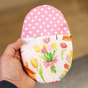 Könnte beinhalten: Ein eiförmiges Stoffobjekt, das in einer Hand gehalten wird, mit einem rosa Tupfenmuster auf der Oberseite und einem Blumendesign mit Tulpen und einer rosa Schleife auf der Unterseite. Wahrscheinlich ein dekoratives Stück oder eine kleine Tasche.