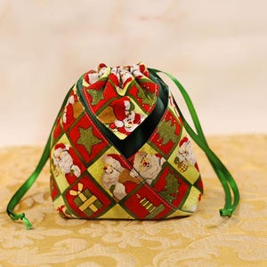 Peut inclure: Une pochette en tissu à cordon de serrage avec un motif à carreaux festif alternant des carrés de Père Noël, d'étoiles vertes, de cadeaux et de bougies. La pochette possède une doublure intérieure vert foncé et des rubans de satin vert.