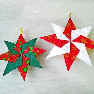 Könnte beinhalten: Zwei handgefertigte sternförmige Weihnachtsschmuckstücke. Eines ist grün und rot mit einem Stechpalmenmuster, das andere ist rot und weiß mit goldenen Schneeflocken-Akzenten. Beide haben goldene Aufhängeschlaufen.