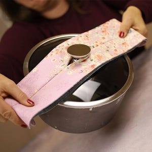 Può includere: Una copertura per manico di pentola con motivo floreale rosa con un bottone in metallo argentato. La copertura è tenuta sopra una pentola in acciaio inossidabile. Il motivo floreale comprende fiori rosa e bianchi su sfondo rosa chiaro.