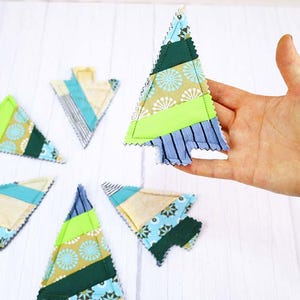 May include: A collection of colorful, handmade Christmas tree ornaments. Each tree is crafted from various fabric scraps in shades of green, blue, and yellow, with a stitched edge. One ornament is held up by a hand.