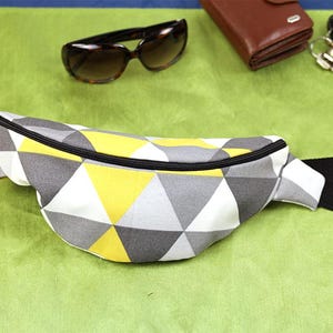 May include: A geometric patterned bum bag in white, grey, and yellow, with a black zip and strap. Sunglasses, a wallet, keys, and a car key are visible in the background. The bum bag is designed to be worn around the waist or across the body.