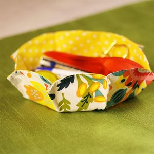 Peut inclure: Un bol en tissu fait à la main avec un motif floral et à pois. Le bol contient des fournitures de couture, dont des ciseaux rouges. Le bol est posé sur une surface verte.
