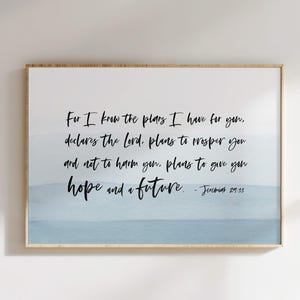 May include: A framed print with a light wood frame. The artwork features a watercolour-style background in shades of blue and white. Black script lettering displays a quote from Jeremiah 29:11, offering a message of hope and future.