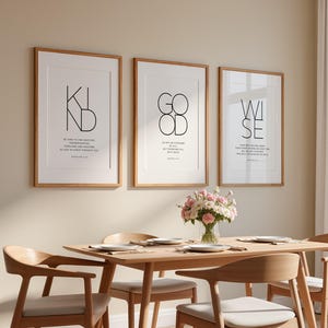 Puede incluir: Tres impresiones artísticas enmarcadas con las palabras "KIND", "GOOD" y "WISE" en fuente negra. Las impresiones están en marcos de madera y cuelgan sobre una mesa de comedor de madera con sillas. Un jarrón de flores rosas está sobre la mesa.