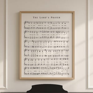 May include: A framed print of sheet music, featuring the text "THE LORD'S PRAYER" at the top. The musical notes are in black on a cream-colored background, encased in a light brown wooden frame. A black piano is in the foreground.