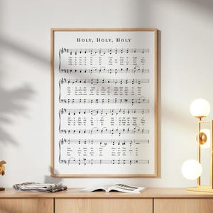 Könnte beinhalten: Ein gerahmter Druck mit Noten, der den Text "HOLY, HOLY, HOLY" trägt. Die Musik ist in Schwarz auf weißem Hintergrund gedruckt und in einem hellen Holzrahmen eingefasst. Eine goldene Skulptur und eine moderne Lampe mit Kugelleuchten stehen auf einem Holzschrank.
