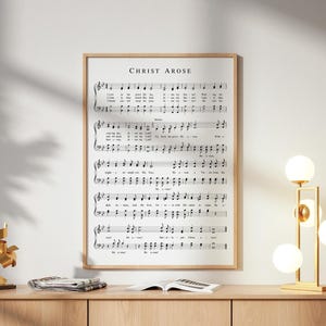 May include: A framed print of sheet music with the text "Christ Arose" at the top. The music is in black ink on a white background, with a wooden frame. The print is displayed above a wooden cabinet.