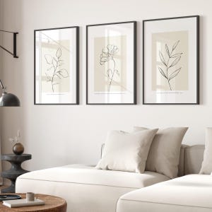 May include: Three minimalist line art prints with black outlines on a beige background. The prints feature a single flower, a branch with leaves, and a branch with leaves. The prints are framed in black and hung on a white wall.